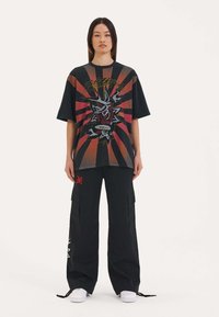 Zwarte t-shirt met rood en oranje grafisch ontwerp met een roos en schedels. Gecombineerd met losse zwarte cargobroeken en witte sneakers.