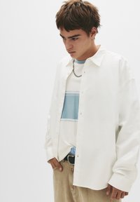 Weißes Hemd mit Knöpfen, über einem hellblauen und weißen gestreiften T-Shirt getragen. Beigefarbene Hose mit einem schwarzen Gürtel und einem blauen gemusterten Detail.