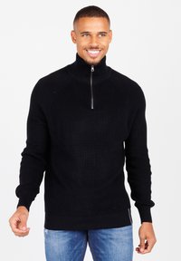Schwarzer Strickpullover mit hohem Kragen und halbem Reißverschluss. Features gerippte Textur und lange Ärmel. Getragen mit blauen Jeans.