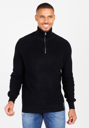 Schwarzer Strickpullover mit hohem Kragen und halbem Reißverschluss. Features gerippte Textur und lange Ärmel. Getragen mit blauen Jeans.