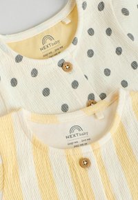 Deux petits tops : l'un avec des pois gris sur un fond crème, et l'autre avec des rayures jaunes et blanches. Les deux présentent des boutons en bois.