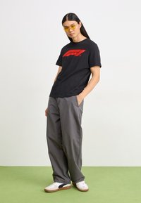 Black cotton t-shirt with a red logo, grey loose-fitting pants, and white sneakers with dark accents, set against a light background.