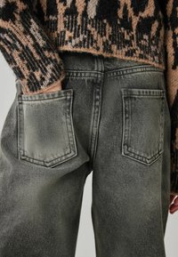 Pantalones de mezclilla verdes con dos bolsillos traseros, presentando una textura desgastada y costuras en contraste, combinados con un suéter de patrón negro y beige.