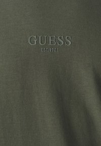 Sötétzöld pamut póló dombornyomott "GUESS" logóval és "EST. 1981" felirattal, sima tapintással és minimalista dizájnnal.