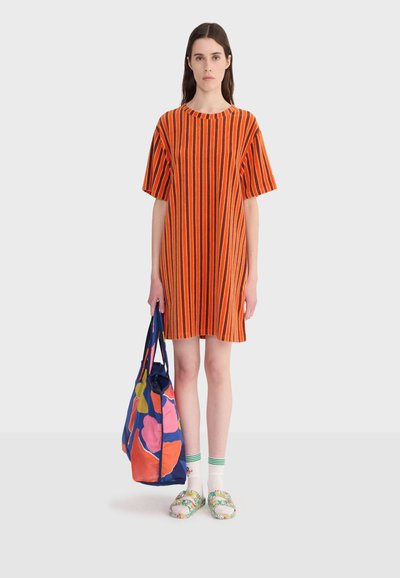Femme debout portant une robe courte rayée orange et noire, des chaussettes blanches, des sandales colorées, tenant un grand sac fourre-tout bleu avec des imprimés floraux abstraits.