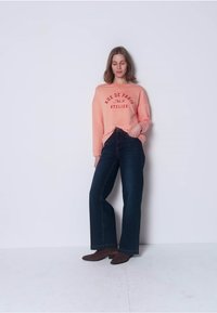 Licht koraalroze sweatshirt met de opdruk "RUE DE PARIS No. 9 ATELIER", gecombineerd met donkerblauwe wijde jeans en bruine enkellaarzen.