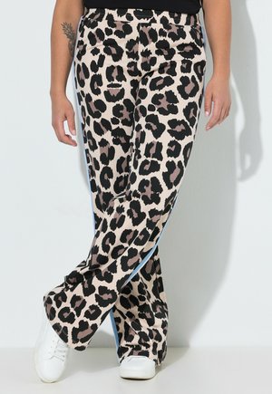 Persona que lleva pantalones con estampado de leopardo en beige, negro y marrón con franjas laterales, zapatillas blancas y un top negro, de pie contra un fondo claro.