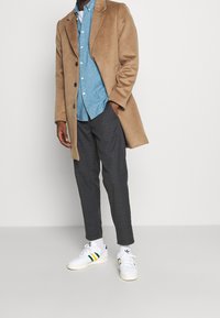 Manteau en laine beige, chemise bleue, pantalon à carreaux gris, baskets blanches avec des rayures vertes et jaunes, présentés sur un fond uni.