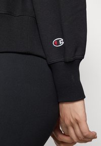 Felpa nera con polsini a costine, dotata di un piccolo logo rosso e bianco sulla manica. Texture liscia, design aderente, stile minimalista.