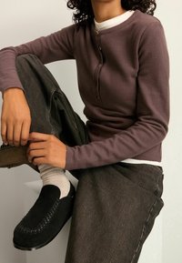 Osoba siedząca z jedną podniesioną nogą, ubrana w brązowy sweter na guziki, ciemne dżinsy, jasne skarpetki i czarne buty wsuwane