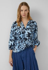 Donna con capelli biondi che indossa una blusa floreale blu con maniche a 3/4 e pantaloni larghi blu scuro, in piedi con una mano nella tasca.