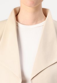 Beige blazer med brede revers og en glat tekstur. Båret over en hvid ribbet top; nærbillede med fokus på udskæringen.