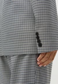 Prim-plan al unei mâini lângă un mânecă de costum cu model pied-de-poule alb-negru și pantaloni asortați, cu patru nasturi negri pe mânecă.