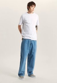 White cotton t-shirt, relaxed fit, paired with loose blue denim jeans. Black and white checkered slip-on shoes. Neutral background.