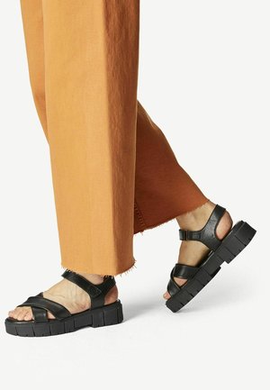 Sandales en cuir noir avec larges brides, semelles épaisses texturées et boucle à la cheville. Portées avec un pantalon large orange présentant des ourlets effilochés.