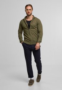Veste zippée vert olive avec capuche, associée à un pantalon noir et des chaussures marron. La veste présente un logo discret sur la poitrine.