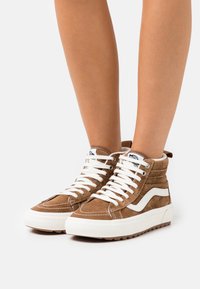 Brune semskede høyhælte sneakers med hvite lisser, kontrasterende hvit side stripe og teksturert gummisåle. Sømdetaljer tilfører interesse.