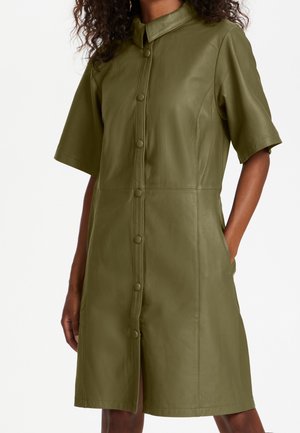 Femme portant une robe en cuir vert olive courte au genou, manches courtes, avec boutons devant et col, debout avec une main dans la poche.