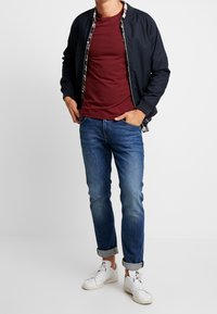 Chaqueta bomber azul marino sobre una camiseta burdeos, combinada con jeans azules con los bajos remangados y zapatillas blancas. La chaqueta tiene un forro de cuadros.