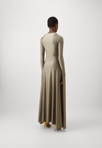 Lang, strakke jurk van lichtbeige stof met lange mouwen en een vloeiende rok, gecombineerd met gele hoge hakken.