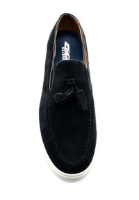 Chaussure slip-on en suede noir avec un bout arrondi, présentant un détail de breloques décoratives et une semelle en caoutchouc blanc. La doublure intérieure affiche un logo de marque.