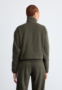 Kort skuren fleecejacka i olivgrön med hög krage och långa ärmar, med en mjuk textur och en ren, minimalistisk design.