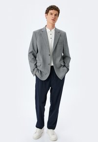 Grauer Blazer mit zwei Vordertaschen, getragen über einem weißen Polo-Shirt. Marineblaue Hose und weiße Sneakers runden das Outfit ab. Glatte Stofftextur.