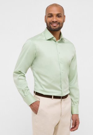 Hellgrüne Hemdbluse aus glattem Stoff mit langen Ärmeln und klassischem Kragen, kombiniert mit beigen Hosen und einem braunen Gürtel.