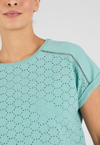 Top en teal claro con un patrón texturizado que presenta agujeros recortados, mangas cortas y un ajuste relajado. Tela suave con un sutil brillo.