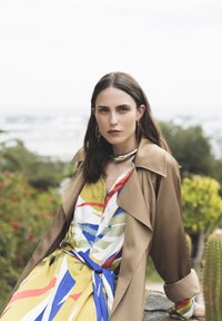 Trench coat di colore marrone chiaro, sovrapposto a un vestito a fantasia colorato con strisce blu, rosse e verdi. Collana a choker metallica lucida particolarmente evidenziata.