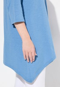 Blaue, kurzärmelige Tunika mit leicht A-Linie-Silhouette und abgerundetem Saum. Weicher, strukturierter Stoff, gezeigt mit einer Hand, die an der Seite ruht.