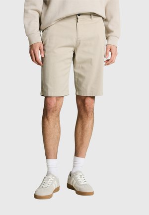 Homme portant un short beige mi-cuisse, des chaussettes blanches montantes et des baskets beiges à semelles en gomme, debout devant un fond uni.