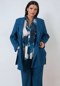 Teal blazer with a relaxed fit, large lapels, and front pockets, layered over a multicolored blouse featuring blue floral patterns.