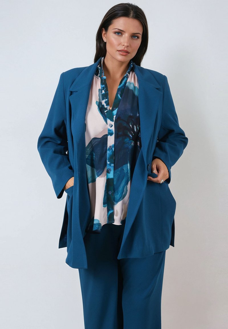Teal blazer with a relaxed fit, large lapels, and front pockets, layered over a multicolored blouse featuring blue floral patterns.