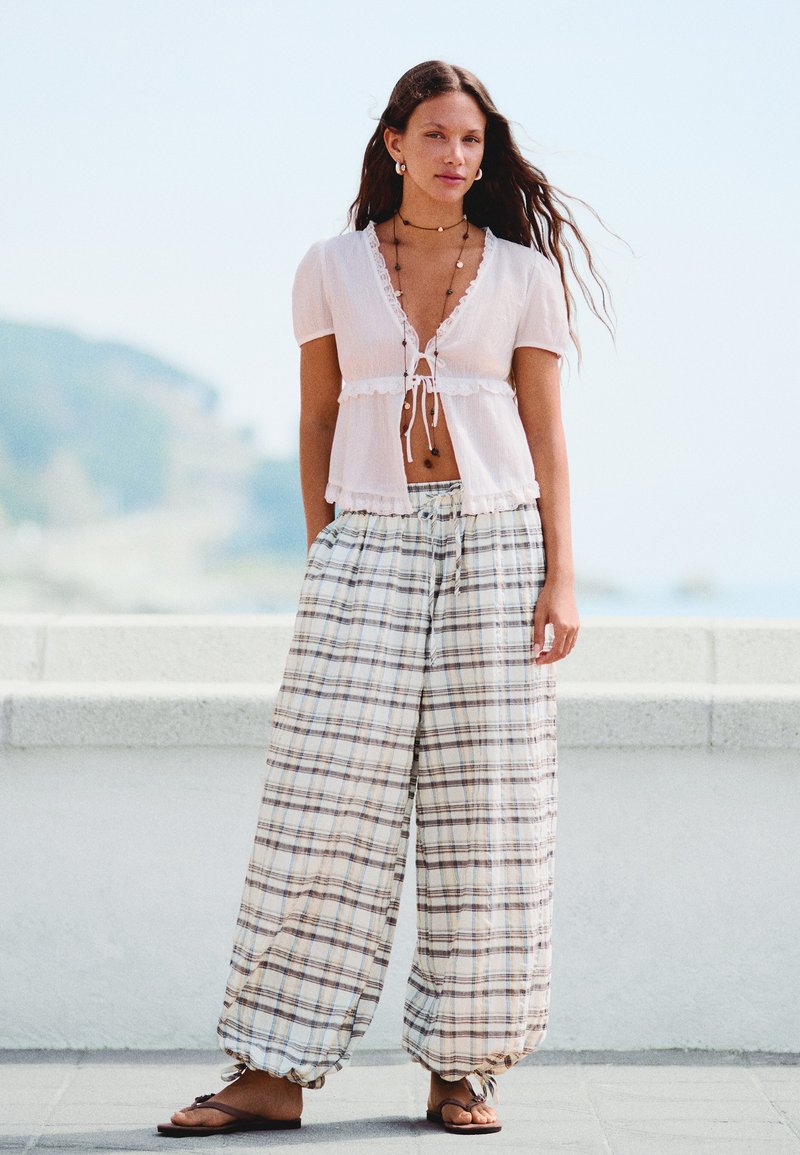 Jeune femme aux longs cheveux portant un chemisier blanc à manches courtes noué à la taille, un pantalon large à carreaux, des sandales et des colliers superposés.