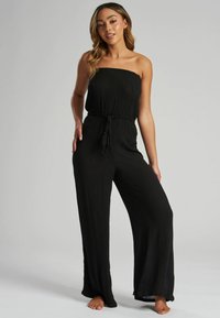 Mono negro sin mangas con un cuerpo ajustado, cinturilla elástica y pantalones de pierna ancha. Hecho de una tela texturizada, presenta un detalle de cordón.