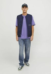 Gilet en matelassé marine par-dessus un t-shirt violet clair, associé à un jean bleu et des baskets noires avec des accents blancs.