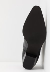 Zapato de cuero negro con punta afilada y tacón grueso. La suela es plana con un acabado texturizado y un sutil detalle del logo.