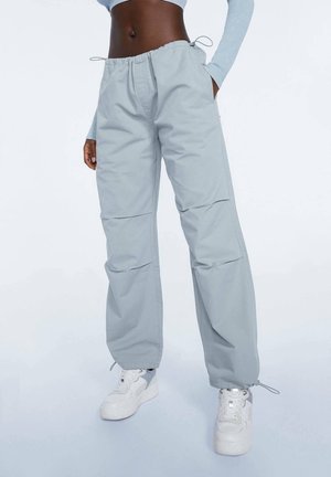 Personne portant un pantalon large bleu clair avec des cordons à la taille et aux chevilles, des baskets blanches et un haut à manches longues court.