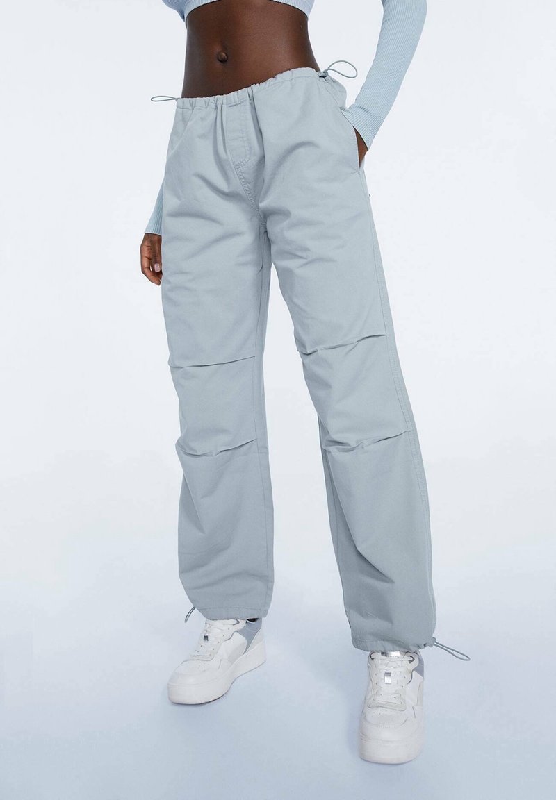Personne portant un pantalon large bleu clair avec des cordons à la taille et aux chevilles, des baskets blanches et un haut à manches longues court.