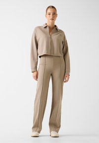 Beżowa skrócona kurtka zapinana na zamek z kołnierzykiem, zestawiona z beżowymi spodniami z wysokim stanem, ozdobionymi pionowymi przeszyciami i szerokimi nogawkami.