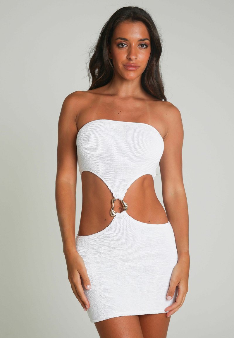 Robe mini tricotée blanche sans bretelles, avec des découpes sur les côtés et un matériel en anneau argenté proéminent à la taille. Tissu lisse et texturé.