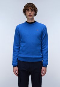 Blå stickad tröja med rund halsringning, ribbade ärmslut och nederkant; har en liten röd logotyp på bröstet. Tyget har en strukturerad yta.