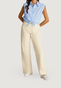 Ljusblå randig kortärmad skjorta knuten i midjan, tillsammans med krämfärgade vida byxor, bärs med vita sneakers.