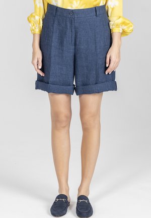 Kobieta ubrana w podwinięte granatowe szorty, żółtą bluzkę w kwiaty z długim rękawem oraz granatowe wsuwane mokasyny, stojąca na jednolitym tle.