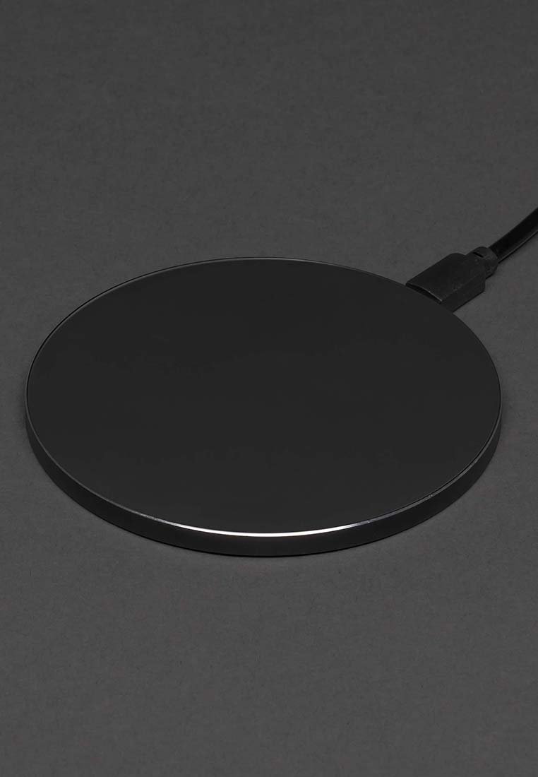 Schwarzes, rundes drahtloses Ladepad mit einem angeschlossenen USB-Kabel auf einer dunkelgrauen Oberfläche.