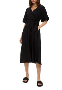 Vestito nero avvolgente in tessuto leggero e testurizzato, con maniche corte, tasche laterali e una cintura da annodare in vita. Indossato con sandali neri piatti.