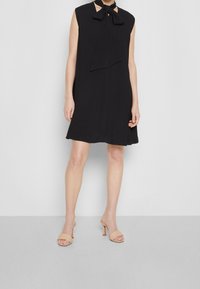 Svart ärmlös klänning med en slipskrage, mjukt tyg, A-linjeformad silhuett som faller ovanför knäna. Matchas med naken öppetåiga klackar.