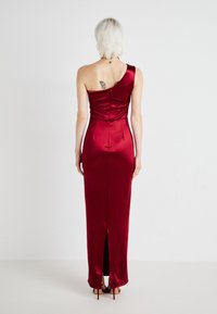 Egy vállra akasztott, testhez simuló estélyi ruha mélyvörös selyemből, oldalsó hasítékkal, sima textúrával és formázott bodice részletekkel.
