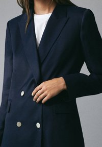 Donna che indossa un blazer doppiopetto blu navy con bottoni argentati su una camicia bianca, mentre aggiusta il blazer con una mano.